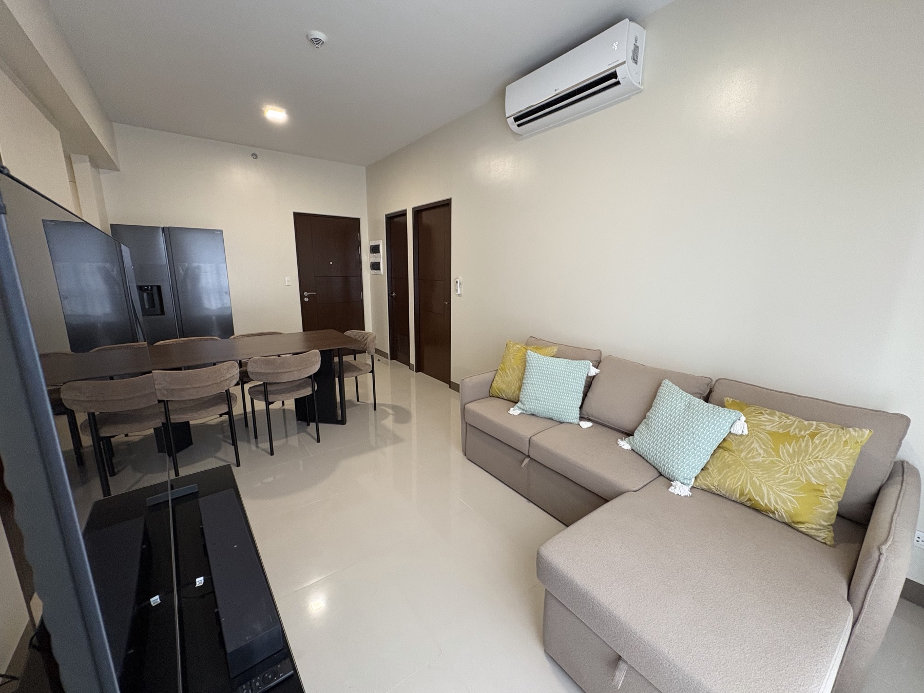Open-plan living and dining room in the DAYON TA two-bedroom condo at Mactan Newtown, with a beige sectional sofa, large smart TV, and tower views through a curtained window